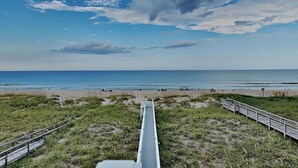 Una playa con un paseo de madera que conduce al agua, zonas verdes y un cielo despejado.