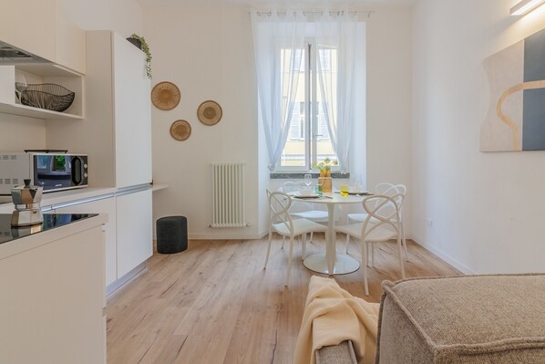 A compact kitchen with white cabinets, a microwave, and a small countertop. There is a dining area with a white table and chairs, and a window with sheer curtains.