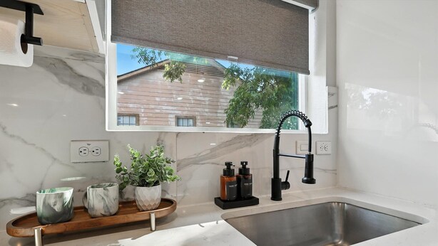 Une cuisine moderne avec un plan de travail en marbre, un évier en acier inoxydable, un plateau en bois avec des tasses, une plante en pot et une fenêtre donnant sur des immeubles.