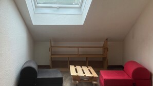 Une petite pièce mansardée avec un puits de lumière, un lit superposé en bois, un canapé rouge, un pouf noir et une table en bois.