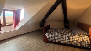 Une chambre avec un plafond en pente, une fenêtre avec des rideaux, un lit recouvert d’une couvre-lit à motifs et un plancher en bois.