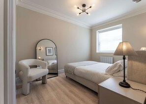 Un dormitorio con cama, una silla, una mesita de noche con lámpara, un espejo y una ventana.