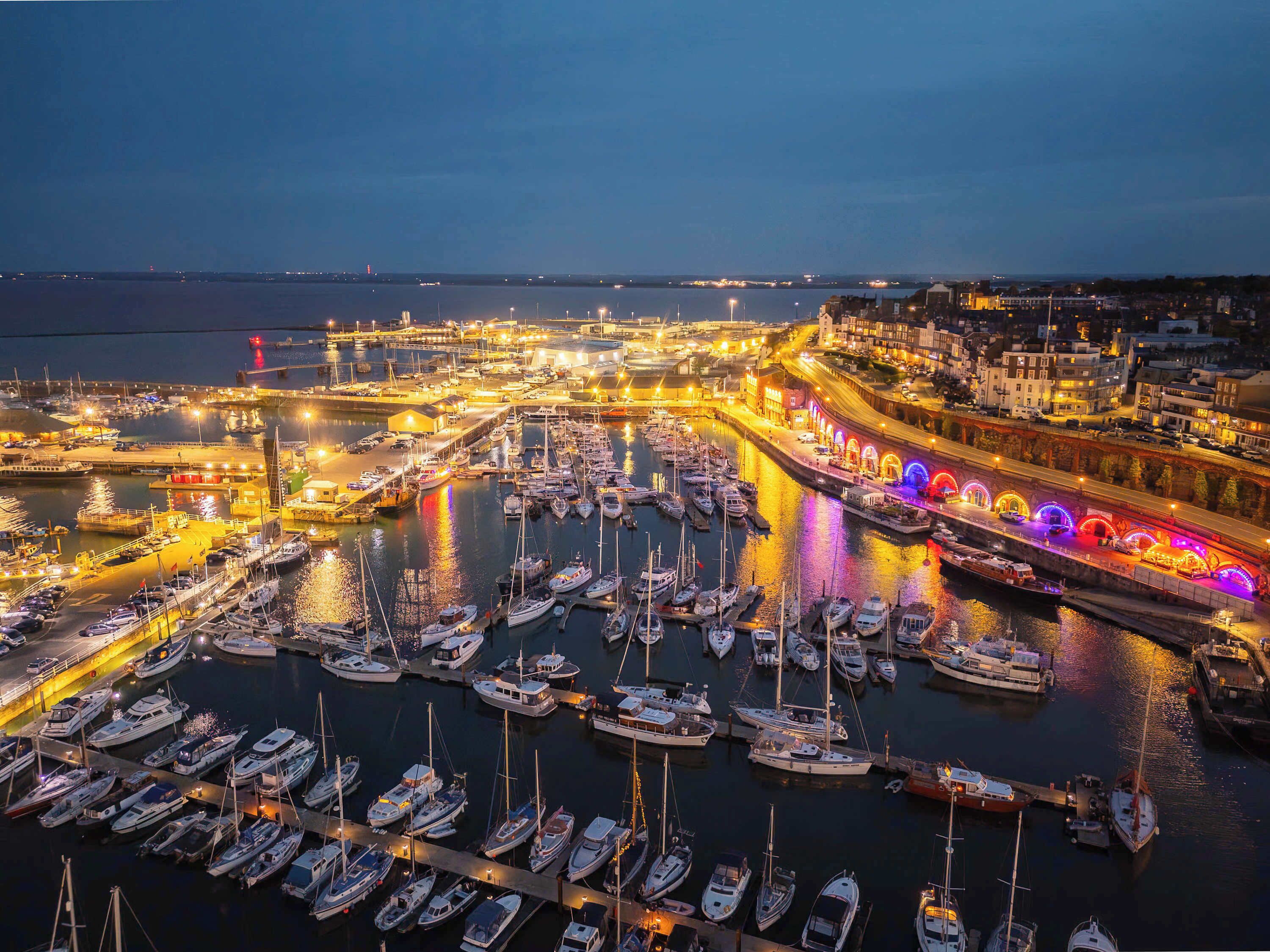 En havn ved skumringen med mange både i havnen, en oplyst promenade og et bybillede i baggrunden.