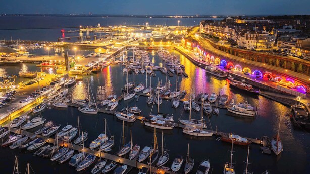 A marina at dusk with numerous boats docked, a brightly lit promenade, and a cityscape in the background.