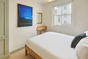 A bedroom with a bed, a window with a view of buildings, a painting on the wall, and a mirror.