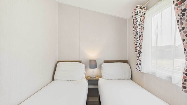 A small, single-room hotel room with two single beds, a nightstand with a lamp, and a window with a polka dot curtain.