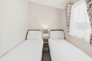 A small, single-room hotel room with two single beds, a nightstand with a lamp, and a window with a polka dot curtain.