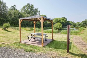 Ein hölzerner Pavillon mit Picknicktischen in einer grünen Parkanlage mit einem Teich im Hintergrund.