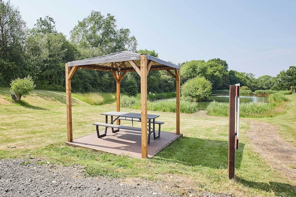 A wooden gazebo with picnic tables in a grassy park area with a pond in the background.