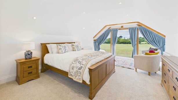 A bedroom with a wooden bed, a nightstand, a dresser, and a view of a garden through a window with curtains. - Host & Stay | Merrow Cottage (Great Malvern)