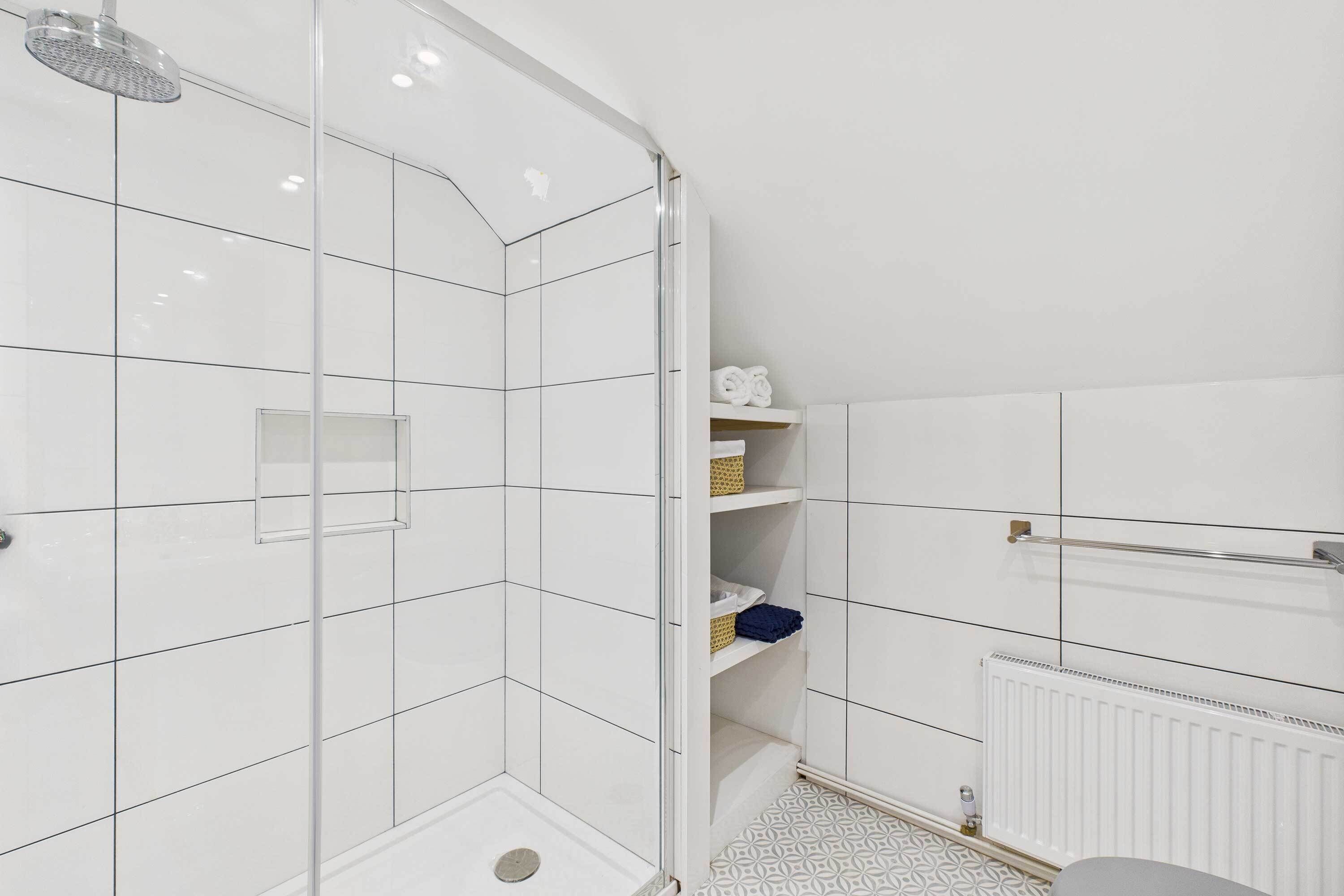 A modern bathroom with white tiles, a glass shower enclosure, a built-in shelf with towels, and a radiator.