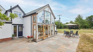 A modern house with a patio area featuring a table and chairs, a large glass door, and a garden. - Host & Stay | Merrow Cottage (Great Malvern)