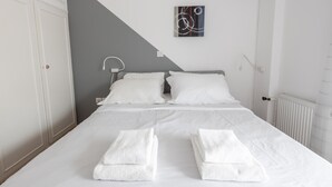 Una cama bien hecha con sábanas y toallas blancas, una pared de acento gris y un cuadro abstracto moderno.