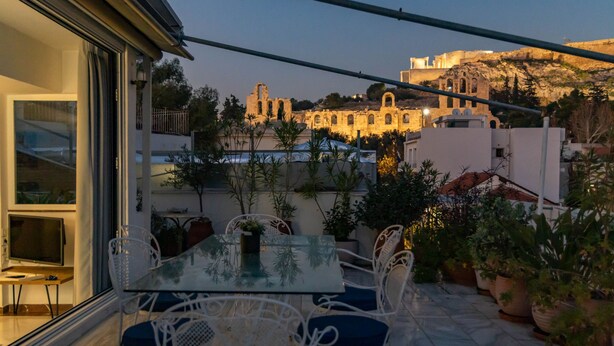Un patio al aire libre con una mesa de vidrio y sillas blancas, con vistas a un edificio histórico y una colina.