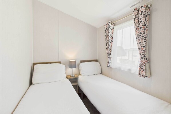 Una habitación pequeña y limpia con dos camas individuales, una mesita de noche con lámpara y una ventana con cortinas a lunares.
