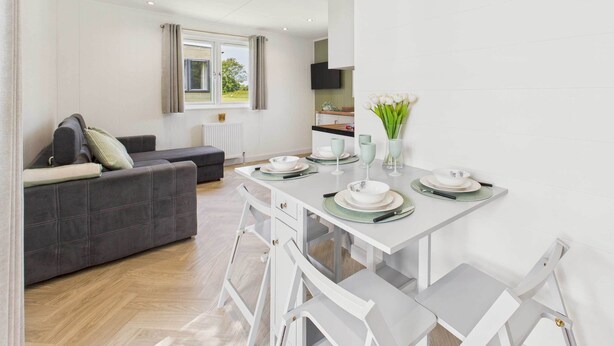 A modern living room with a grey sofa, a white dining table set with plates and glasses, and a kitchen area in the background.