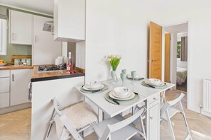 Eine moderne Küche mit weißen Schränken, einem Esstisch gedeckt mit Tellern und Gläsern und Blick in ein Schlafzimmer durch eine offene Tür.