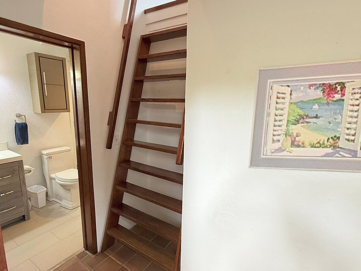 Eine Treppe mit Holzbodenstufen führt zu einer Zwischenebene, an einer weißen Wand hängt ein Bild und es gibt ein Badezimmer mit Toilette, Waschbecken und Schrank.