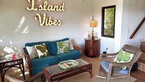 A cozy living room with a teal sofa, wicker furniture, and a wall sign reading "Island Vibes". - Charming Corner Condo with A/C, Stunning Water Views, and Gorgeous Sunsets! (St John)