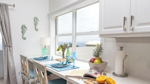 A kitchen with a window, a table set with plates, a bowl of fruit, and a cutting board with a knife. - Sunset Dream - 1BR Hillside Retreat – A/C & Ocean Views, Newly Updated. (St John)