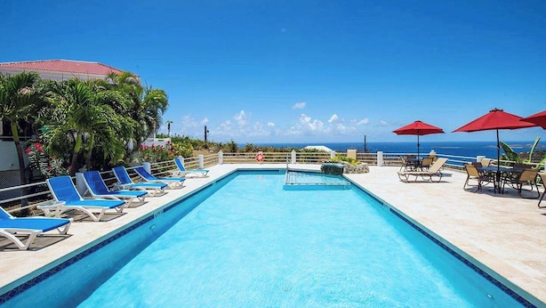 A swimming pool with blue lounge chairs and red umbrellas, surrounded by palm trees and a view of the ocean. - Sunset Dream - 1BR Hillside Retreat – A/C & Ocean Views, Newly Updated. (St John)