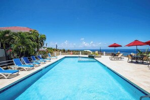 A swimming pool with blue lounge chairs and red umbrellas, surrounded by palm trees and a view of the ocean. - Sunset Dream - 1BR Hillside Retreat – A/C & Ocean Views, Newly Updated. (St John)