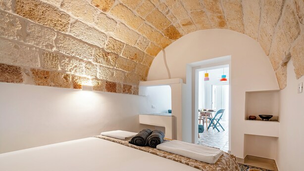 A hotel room with a stone wall, a bed with white linens, a small table with a bowl, and a view of the outside. - Pescamare Apartment Polignano A Mare (Polignano a Mare)
