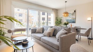 Una sala moderna con sofá, mesa de centro y sillas, una mesa de comedor con sillas, un ventanal con vista a edificios y una lámpara colgante.