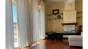 A spacious room with a wooden floor, a sofa, a fireplace with a painting, and a view through large windows with curtains. - 5 min from the sea large Apartment - Wifi and Air Conditioning (Lido di Camaiore)