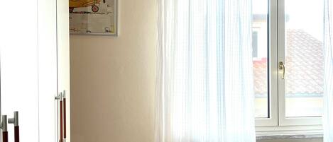 Une chambre avec une fenêtre, un radiateur, un lit avec du linge de lit blanc et un tableau accroché au mur.