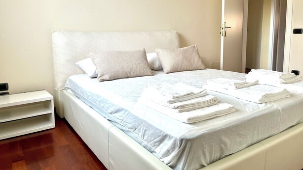 A neatly made bed with pillows and folded towels, a bedside table, and a door leading to another room. - 5 min from the sea large Apartment - Wifi and Air Conditioning (Lido di Camaiore)