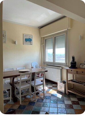 Een klein eetgedeelte met een tafel, stoelen, een radiator, een raam met zonwering en een bureau met een lamp en een fotolijst.