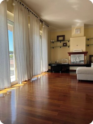Amplia habitación con suelo de madera, sofá, chimenea con un cuadro y vistas a través de amplias ventanas con cortinas.