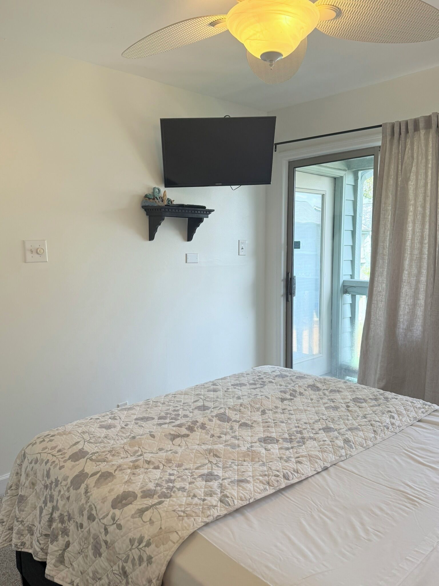 A bedroom with a bed, a ceiling fan, a mounted TV, and a window with curtains.