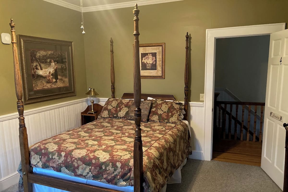 Una cama con dosel con ropa de cama floral, una mesita de noche con lámpara y dos cuadros en la pared.