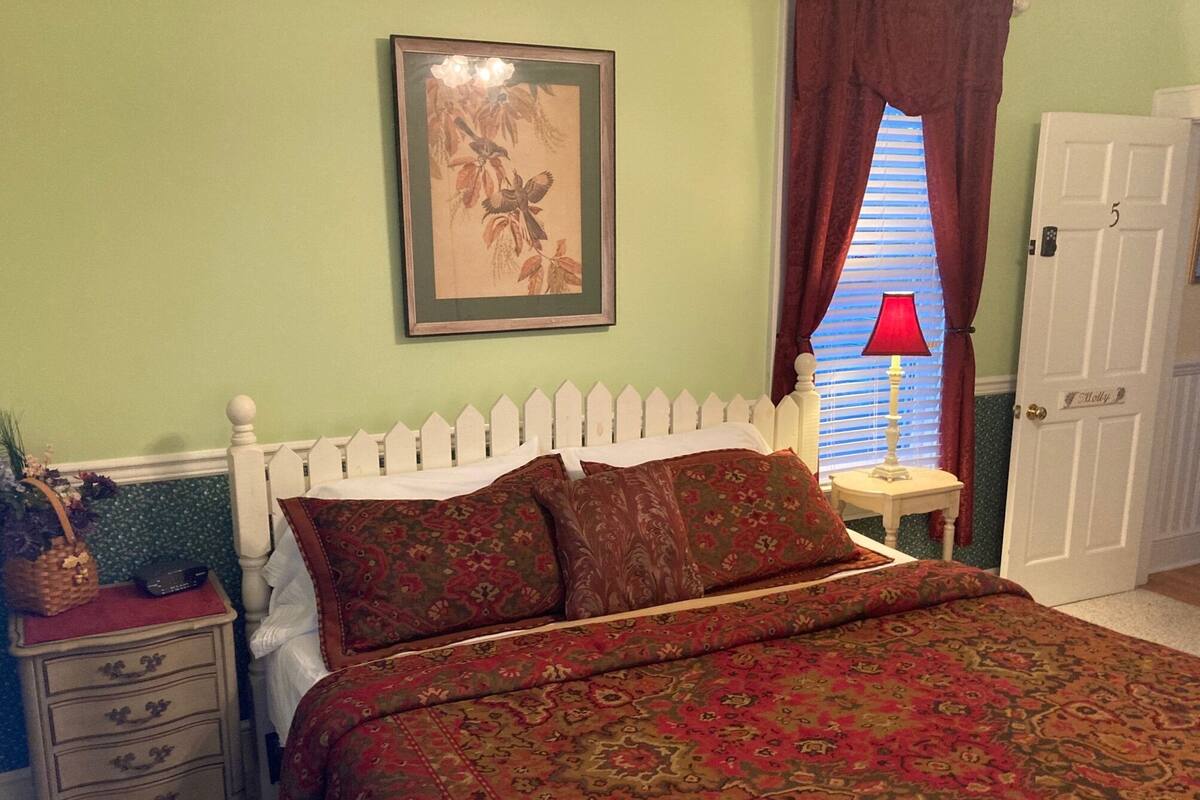 Un dormitorio con estructura de cama blanca, colcha estampada, mesita de noche con lámpara y una puerta con la etiqueta 'Molly'.