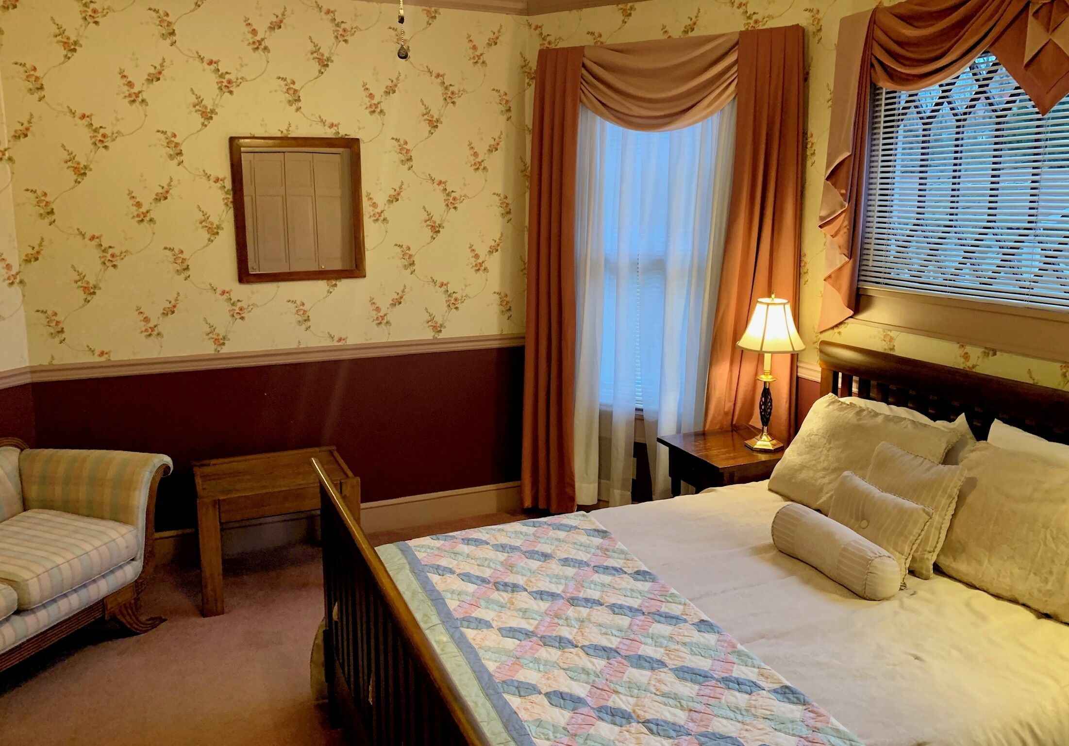 A hotel room with a bed, a nightstand with a lamp, a chair, and a window with curtains.