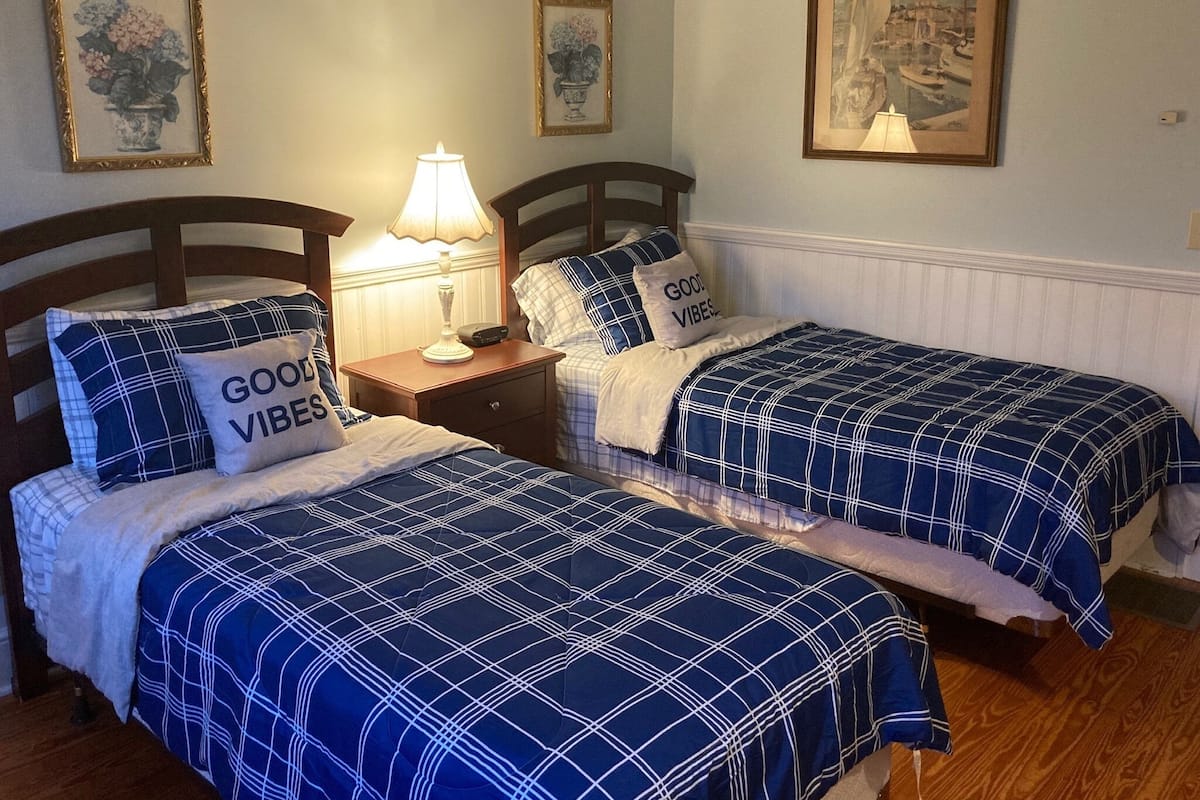 Un dormitorio con dos camas, cada una con colcha y almohadas a cuadros azules y blancos con el texto “GOOD VIBES”. Entre las camas hay una mesita de noche con una lámpara, y en la pared sobre la mesita de noche, un cuadro enmarcado.