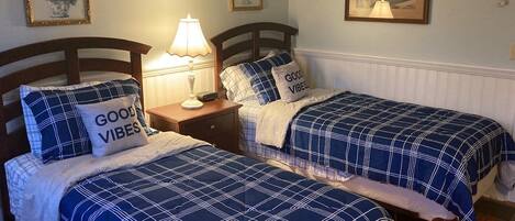 A bedroom with two beds, each with a blue and white checkered bedspread and pillows with the text "GOOD VIBES". There is a nightstand with a lamp between the beds, and a framed picture on the wall above the nightstand.