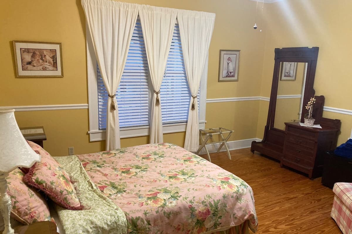Un dormitorio con una colcha de flores, un aparador de madera, un espejo y una ventana con cortinas.