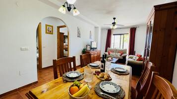 Un comedor con una mesa de madera dispuesta para una comida, un sofá al fondo y un ventilador de techo.
