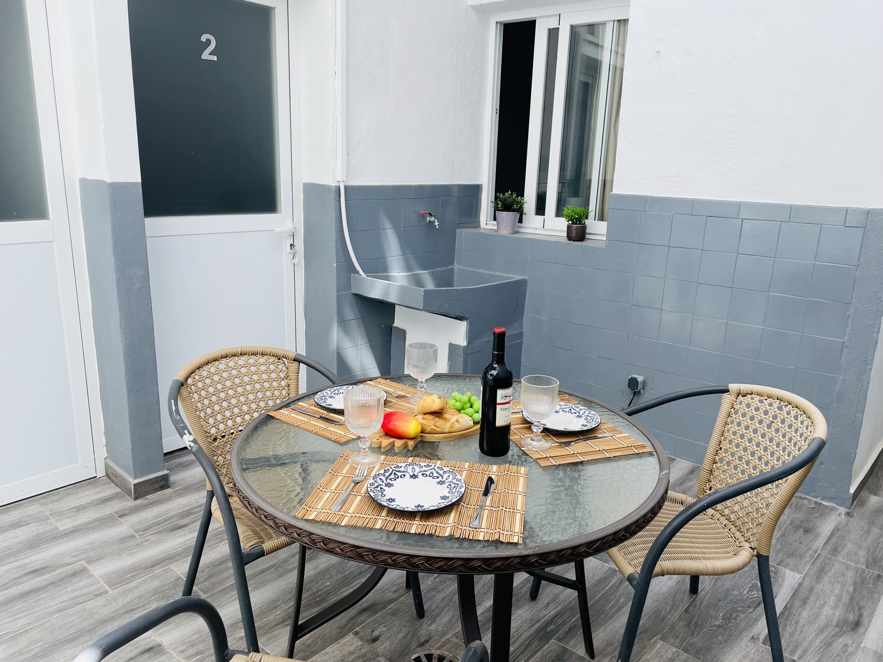 A round glass table with four wicker chairs, a bottle of wine, and a fruit bowl.