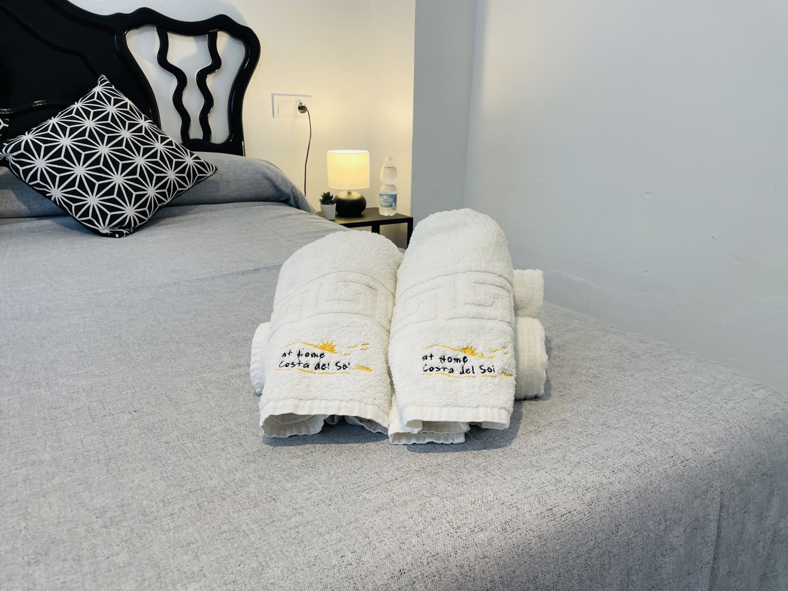 A bed with a geometric patterned pillow, a white towel with text, and a bedside table with a lamp.