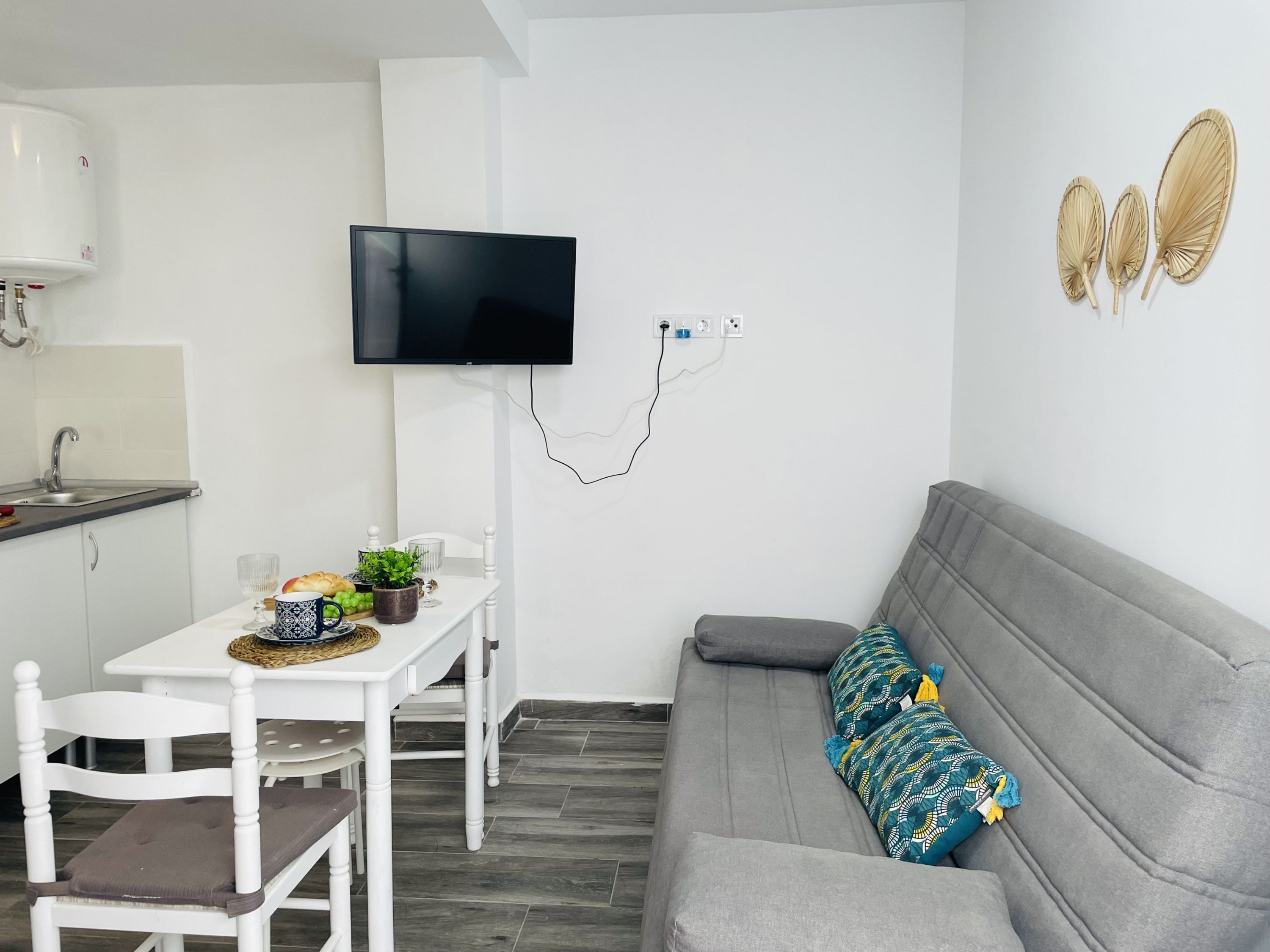 A modern living space with a grey sofa, a white dining table with chairs, a flat-screen TV mounted on the wall, and a small kitchen area with a sink and a water heater.