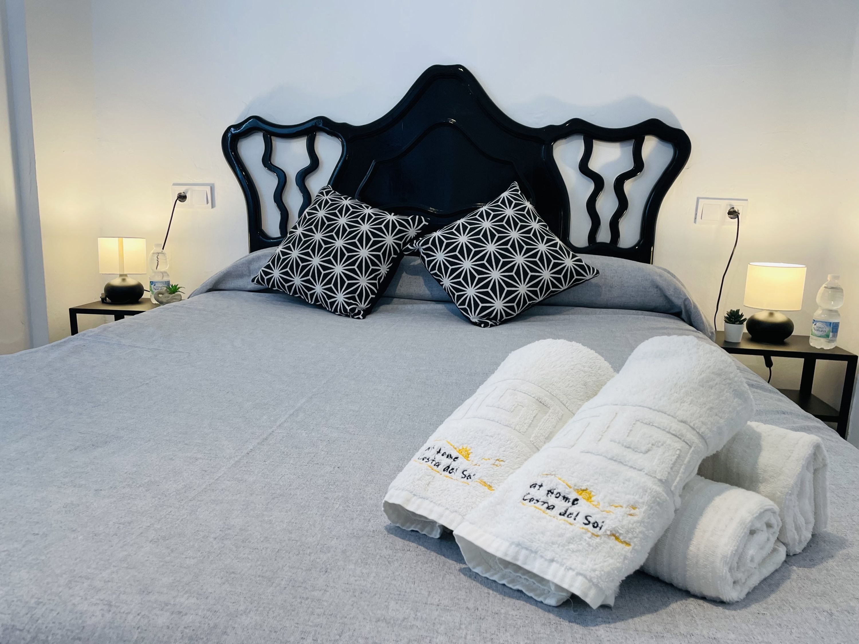 A neatly made bed with geometric patterned pillows, a headboard, and rolled white towels with text.