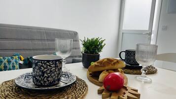 Un coin repas avec une table blanche, deux tasses à motifs, un set de table en fibres tressées, un pain, une tomate rouge et une plante en pot.