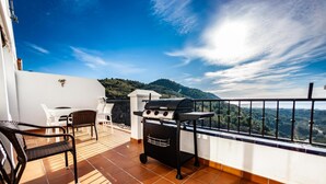 A balcony with a grill, a table, and chairs, overlooking a mountainous landscape.