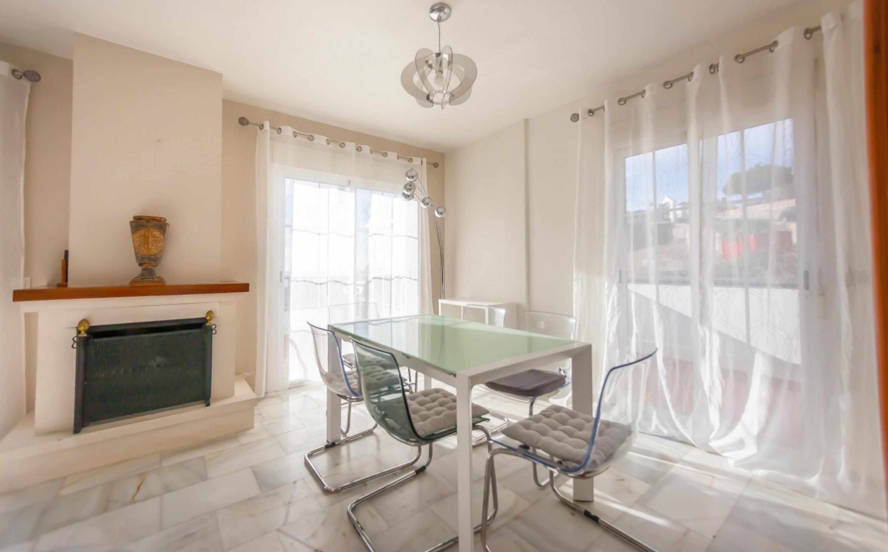 A dining area with a glass table and chairs, a fireplace, and large windows with sheer curtains.
