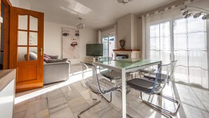 A modern living room with a glass dining table, metal chairs, a sofa, and a TV.
