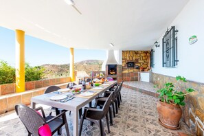 An outdoor dining area with a stone-tiled floor, a long table set for a meal, and a stone fireplace. - Villa Sanchez Sol by At Home Costa del Sol (Torrox)
